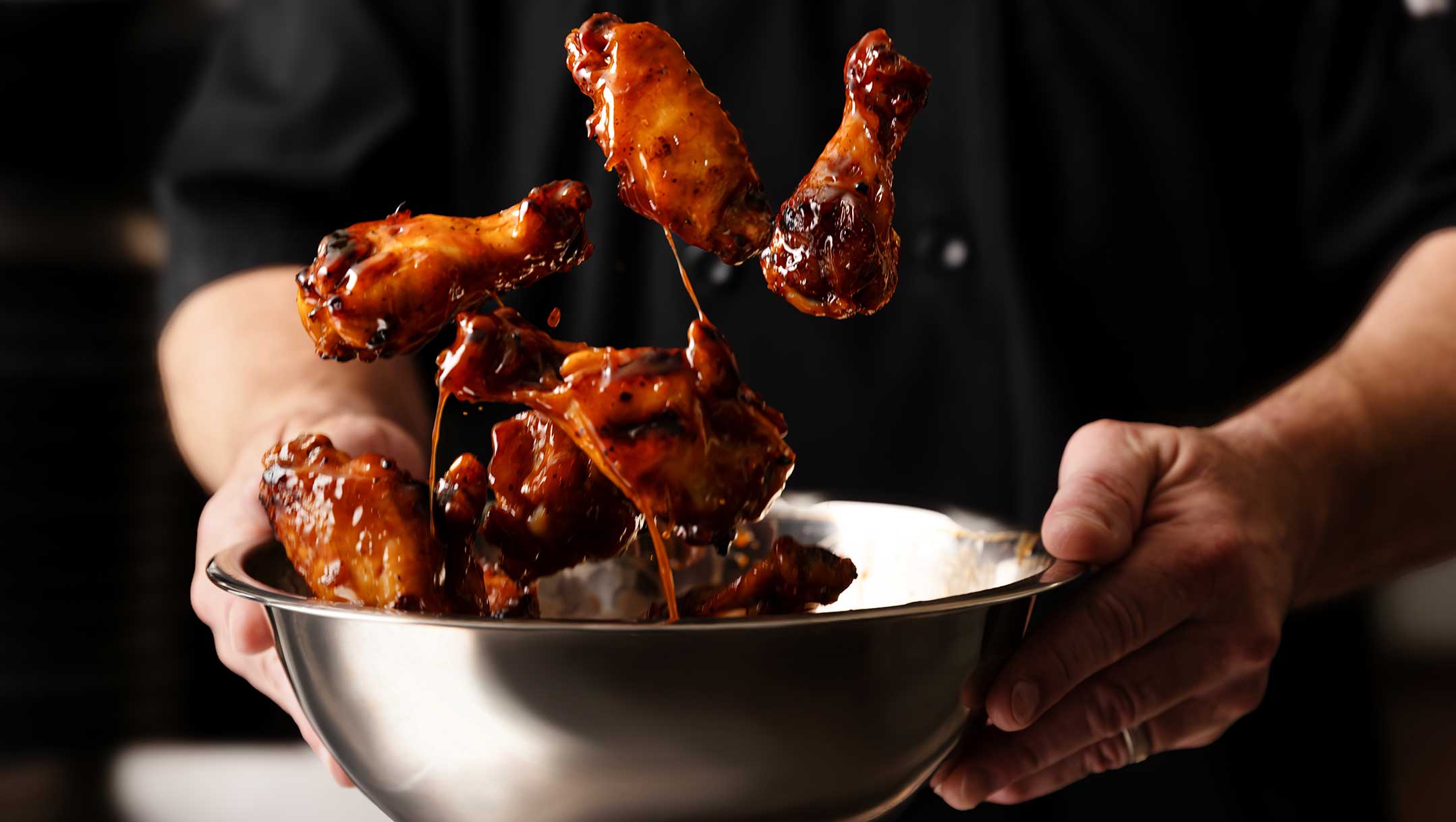 Wings being sauced and tossed in a bowl by the Chef in a black coat