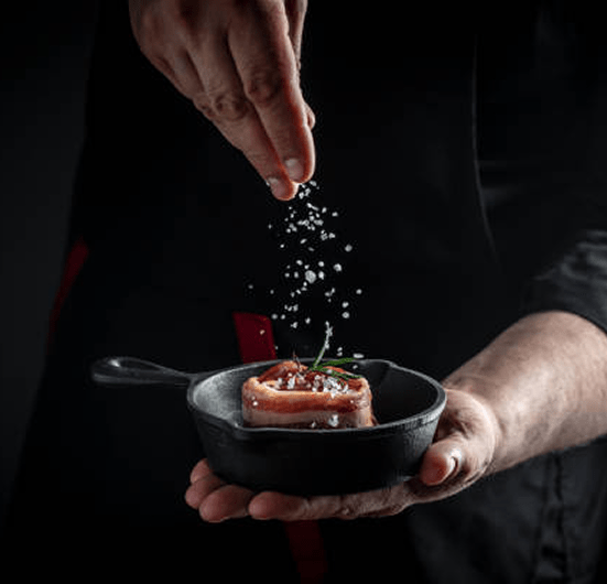 Chef sprinkling coarse sea salt over a filet wrapped in bacon in a cast-iron skillet, symbolizing creativity and attention to detail in restaurant marketing.