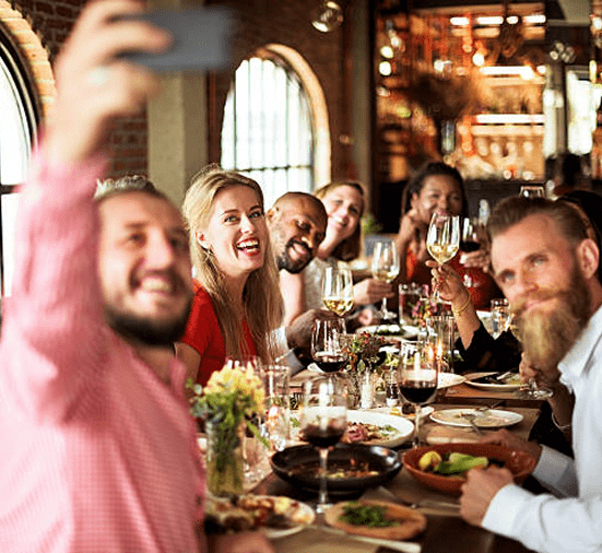 Group of friends taking a selfie while enjoying food, wine and conversation at a lively restaurant table.