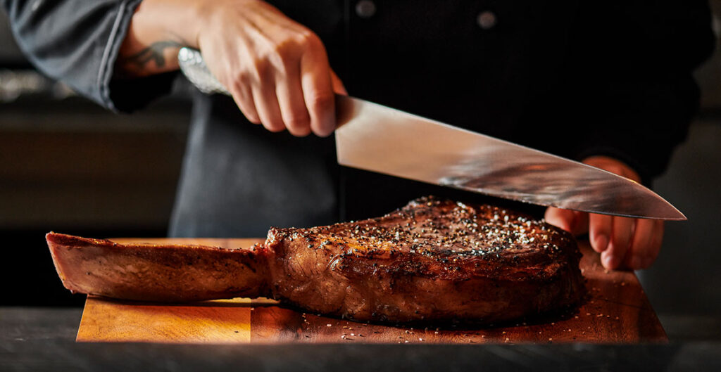 Fleming's Tomahawk about to be sliced with a large knife by a chef in a black coat