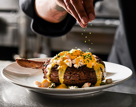 Fleming's Chef sprinkling parsley on the Bone-In Ribeye with Lump Crab and oscar-style sauce